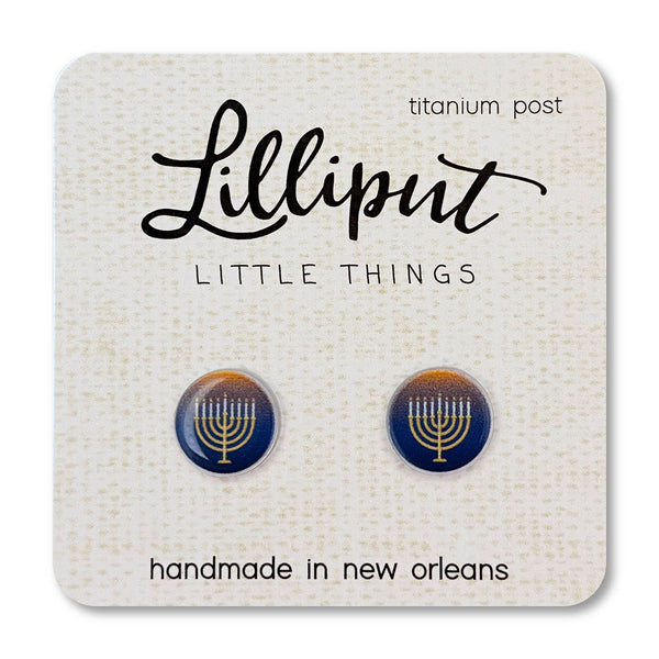 A pair of round stud earrings displayed on a textured cream card. The earrings have a dark blue base with a gold menorah symbol in the center and a gold ring around the edge.